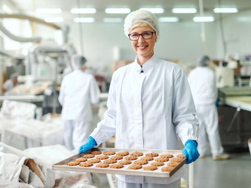 A woman wearing a white hairnet and gloves is presenting cookies made in a food workshop.
