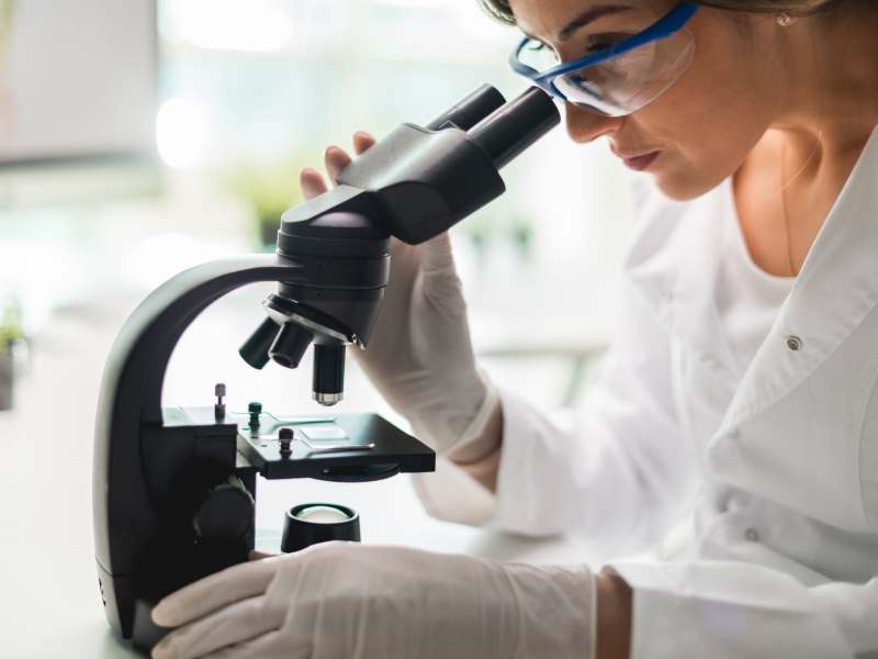 A woman wearing gloves is looking into a microscope.