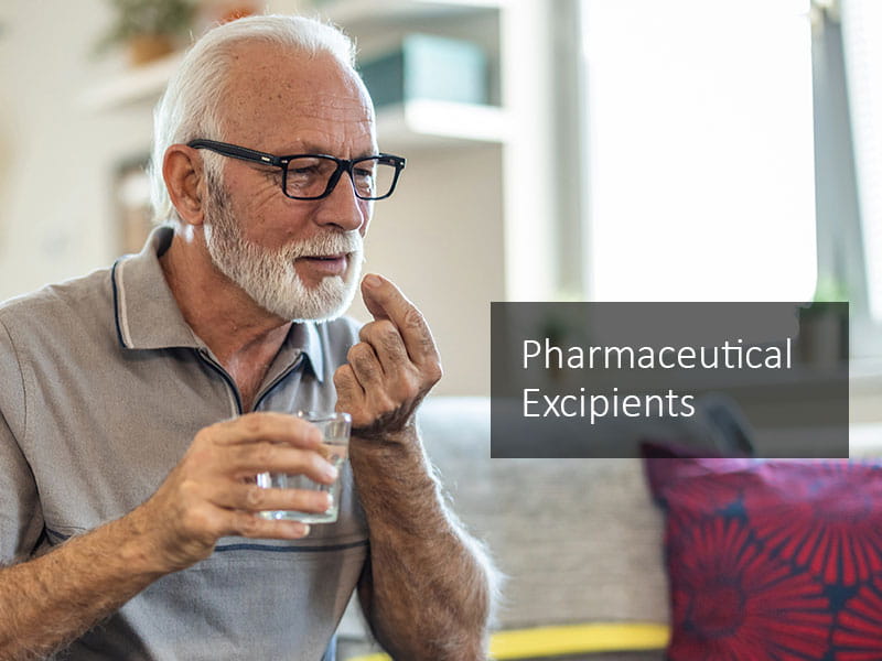 Older man with short white hair and beard, wearing glasses, taking a pharmaceutical tablet with a glass of water.