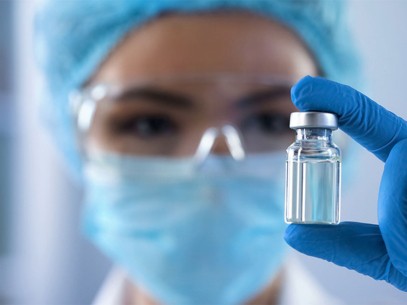 Gloved hand holding a small glass vial in a laboratory setting with medical attire in the background.