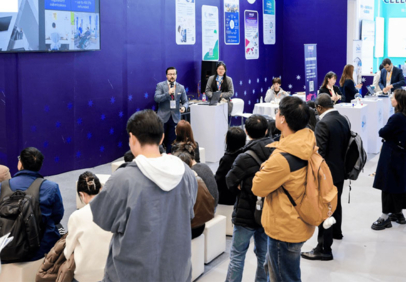 An audience at a professional trade show, with several people attending and presenters speaking or sitting at booths.