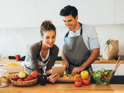 Two people cooking together in a kitchen with fresh vegetables, fruits, and a salad on the counter.