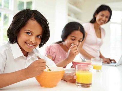 Children eating breakfast with bowls and orange juice on a kitchen table, adult in the background.