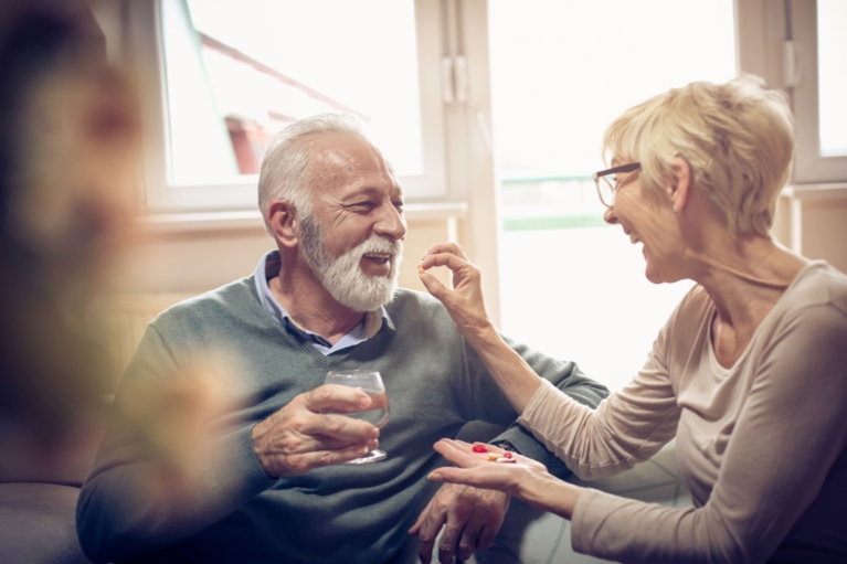 Older couple smiling as the woman administers a red tablet to the man.