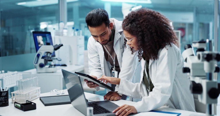 Two scientists in a laboratory wearing white lab coats reviewing data on a tablet and laptop.