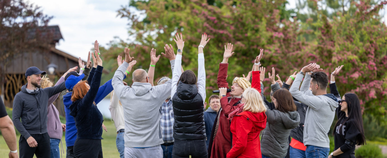 Employees at a team-building activity, standing in a circle, raising their hands to the sky, with green trees and a wooden building in the background.