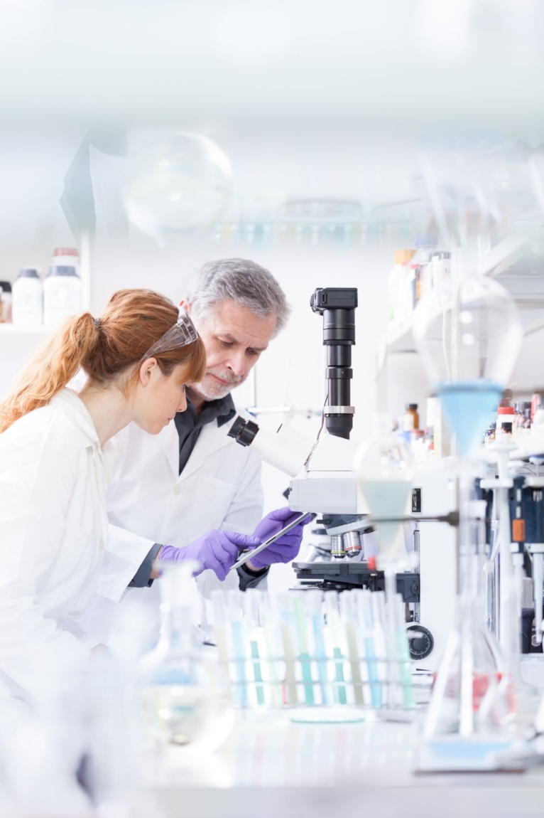 Two scientists in a laboratory wearing white lab coats reviewing data on a tablet and laptop.