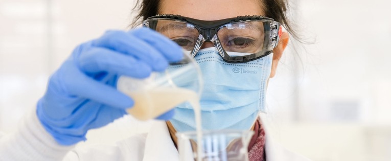 Femme scientifique portant des lunettes de sécurité, une blouse blanche et des gants bleus, versant un liquide dans un récipient dans un laboratoire lumineux.