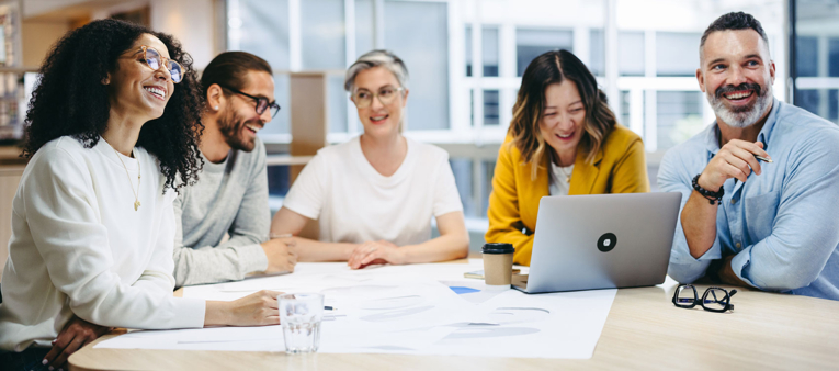 Cinq personnes souriantes sont réunies autour d'une table, des documents et un ordinateur portable posés devant eux.