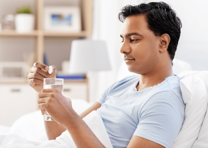 A man sitting in bed with a glass of water holding a tablet